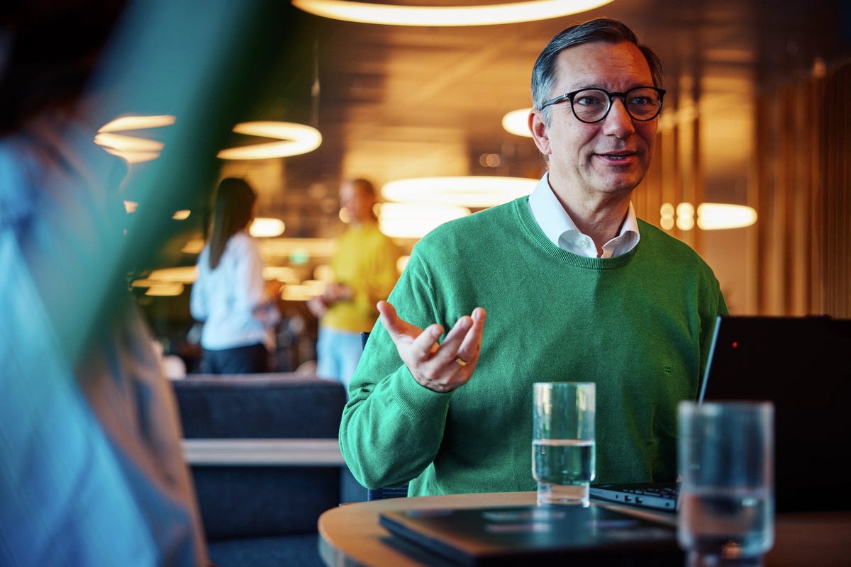 Man in a green sweater sitting at a table with a water glass, engaged in conversation.