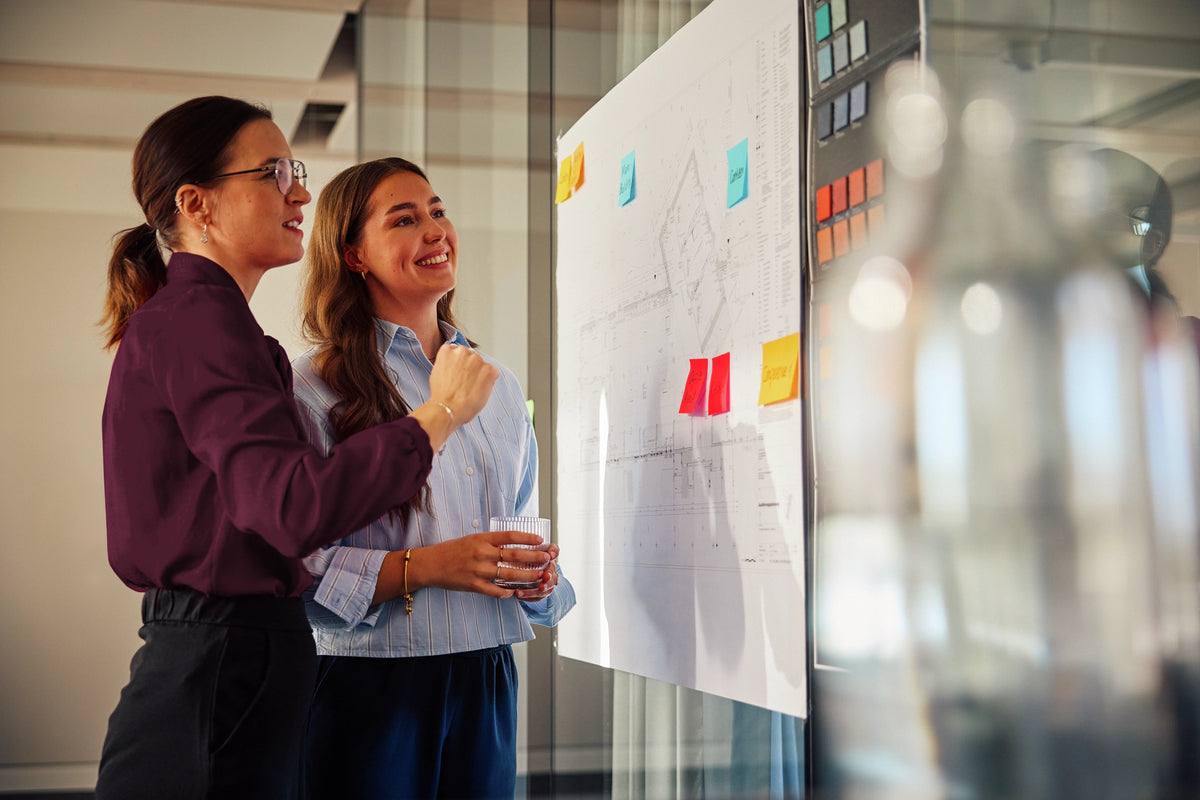 Two women looking at a whiteboard with colorful sticky notes in an office setting.