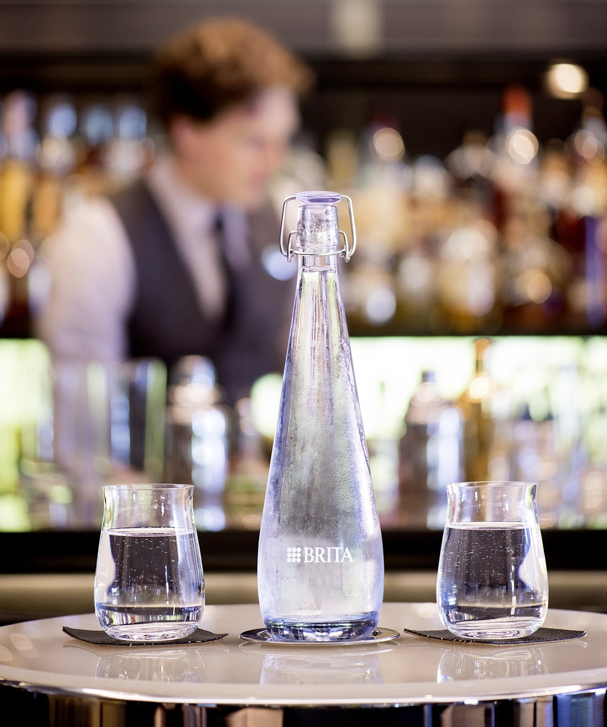 Bouteille Swing  BRITA Bouteille Swing  accompagnée de deux verres d'eau posés sur le comptoir d'un bar, avec un barman flou en arrière-plan.