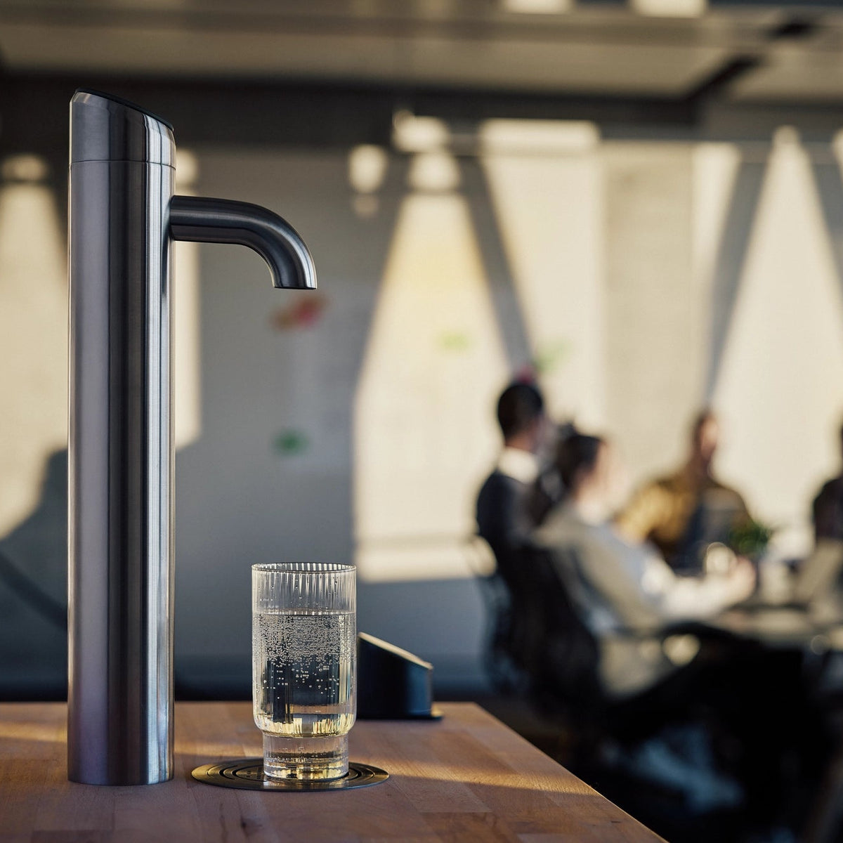 fontaines à eau  BRITA Extra I-Tap posée sur une table en bois, avec un verre d'eau placé en dessous, dans un décor de bureau flou.
