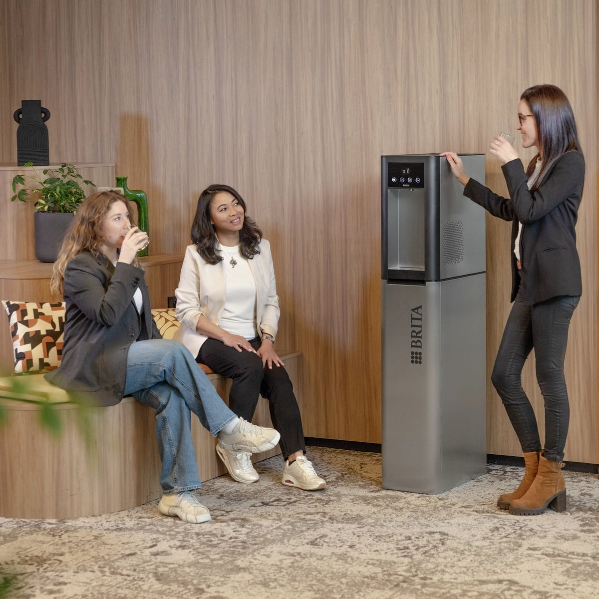 Une femme debout à côté d'un fontaines à eau  Top Pro dans un bureau moderne, en compagnie de deux autres femmes assises sur un banc.