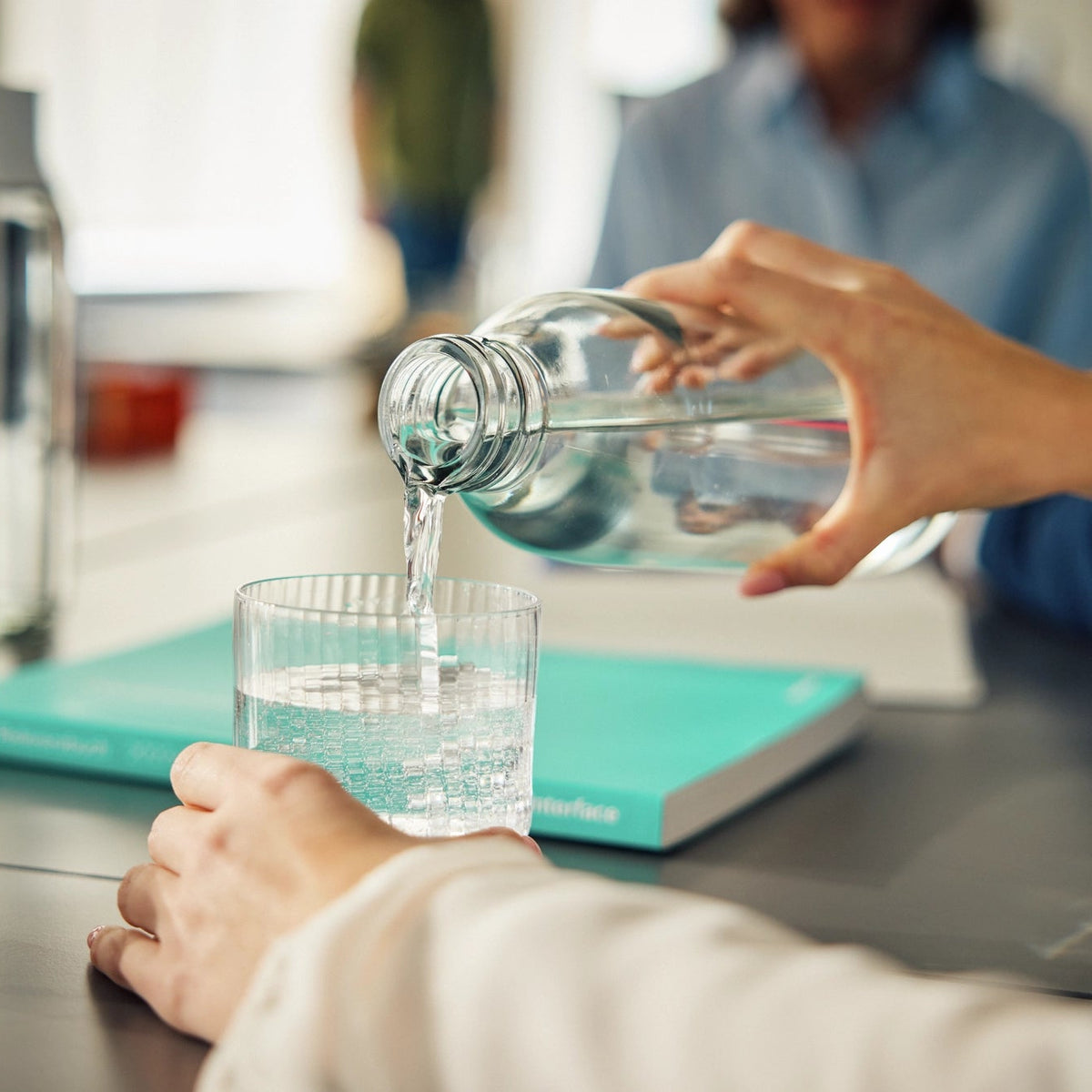Une personne en train de verser de l'eau d'une bouteille dans un verre posé sur un bureau.